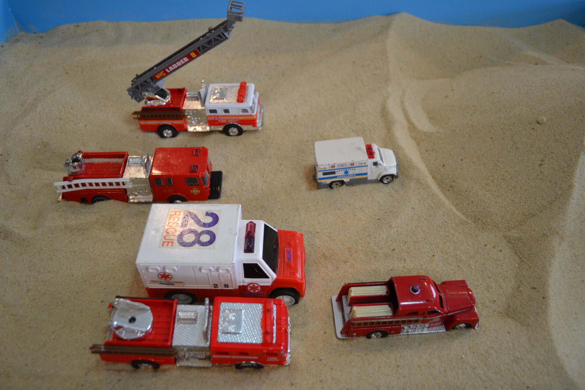 A group of toy cars on sand with fire truck and ambulance.